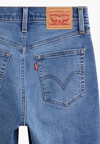 Jean en denim bleu clair avec une patch en cuir à la taille, des poches arrière cousues avec un design distinctif en V, et des coutures contrastées.
