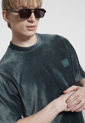 Giovane uomo con occhiali da sole scuri e camicia a maniche corte grigio scuro con un piccolo logo "Hugo" sul petto, mani intrecciate.