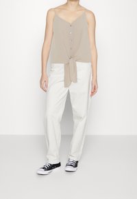 Mujer vestida con una blusa sin mangas de color beige, con botones y lazo en la parte frontal, pantalones blancos de pierna recta y zapatillas negras de corte bajo, de pie contra un fondo blanco.