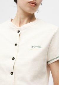 Κοντινό πλάνο ενός ατόμου που φοράει μια κρεμ πουκάμισο Columbia με μαύρα κουμπιά και πράσινη επένδυση στο κοντό μανίκι.