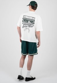 Jeune homme portant un t-shirt blanc avec une illustration et un texte sur un café, un short vert foncé, une casquette assortie, des chaussettes blanches et des mocassins noirs, debout devant un fond gris.