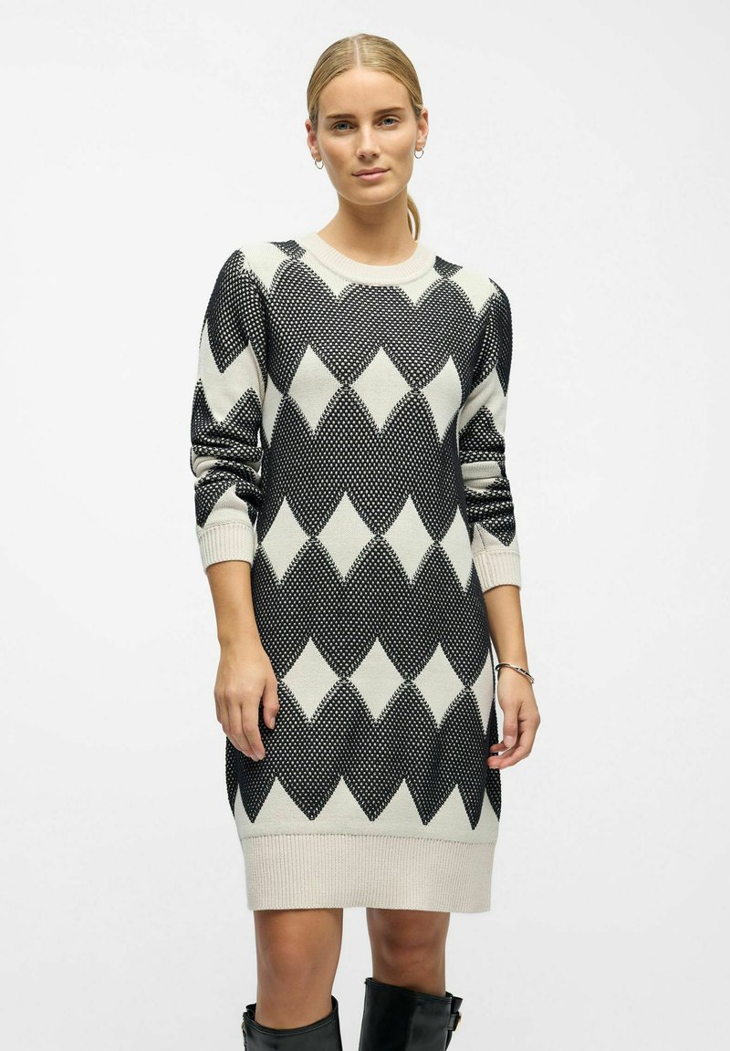 Robe en tricot noire et blanc cassé avec un motif en diamant. Elle possède un col rond et des poignets côtelés. Mi-longue avec une coupe décontractée.