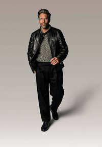 Veste en cuir noir sur une chemise à motif, pantalon noir et baskets noires. Détails notables : lunettes et design texturé de la chemise.