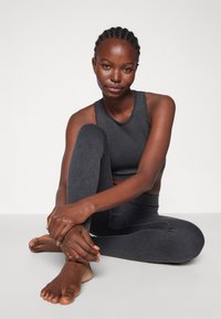 Juoda raukšlėta sportinė maikutė ir suderinti aukšto liemens leggingai su tekstūruotu raštu. Modelis sėdi su suglaustomis rankomis ir basomis kojomis.