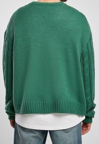Pull vert surdimensionné en maille avec détails torsadés sur les manches, ourlet et poignets côtelés, et col rond. Porté par-dessus une chemise blanche.
