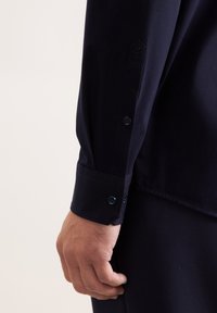 Camicia blu scuro con polsini bottonati a due bottoni, tessuto strutturato e un dettaglio esagonale vicino alla manica.