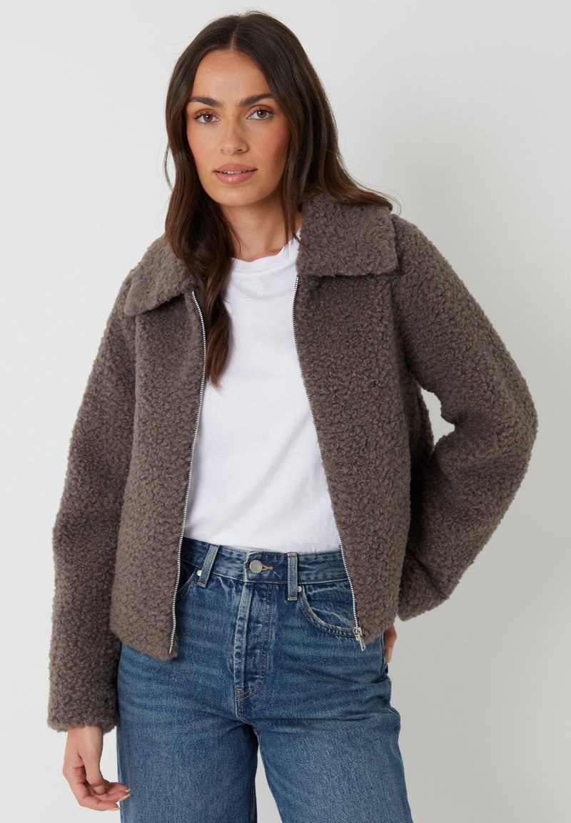 Brun, fluffy zip-up jakke med spredt krave, tekstureret stof og lange ærmer, parret med en hvid T-shirt og high-waisted blå jeans.