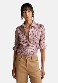Hemd mit Knöpfen in einem geometrischen Muster in Pink und Lila auf einem cremigen Hintergrund. Aufgerollte Ärmel und ein kragenloser Ausschnitt. Trägt beigen Hosen.