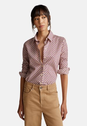 Een button-up shirt met een geometrisch patroon in roze en paars op een crèmekleurige achtergrond. Opgerolde mouwen en een kraag. Draag met beige broek.