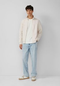 Lichtbeige ritsjack over een wit shirt, gecombineerd met lichtblauwe losse jeans en beige sneakers met witte accenten. Eenvoudig ontwerp, zachte textuur.