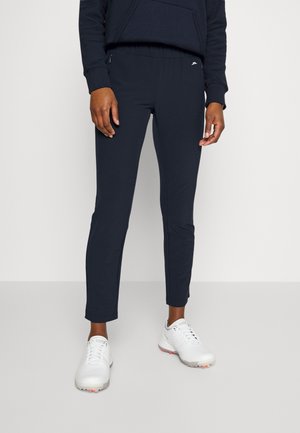 Persoon met zwarte slim-fit broek en witte sportschoenen, staand tegen een effen lichte achtergrond.