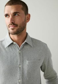 Pilkas marškinis su mygtukais, pagamintas iš minkštos medžiagos, turintis smailų apykaklę, krūtinės kišenę ir tekstūrinį mezginio raštą.