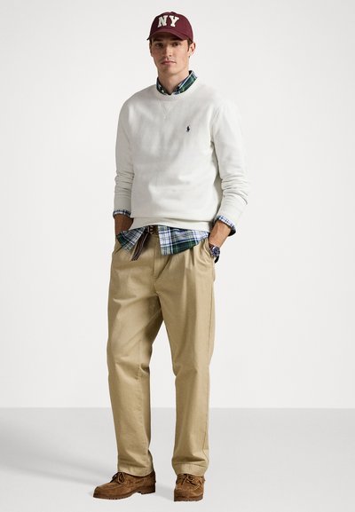Jonge man met een bordeauxrode NY pet, witte trui over een groene geruite blouse, beige broek, bruine schoenen, staand met handen in de zakken.
