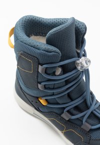 Stivale blu alto con mesh e texture lisce, caratterizzato da accenti gialli, un sistema di bloccaggio trasparente per i lacci e dettagli di cucitura a contrasto.