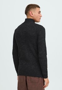 Mørk grå strikkegenser med høy krage, ribbestrikkede mansjetter og kant. Stoffet har en teksturert overflate, designet for en tettsittende silhuett.