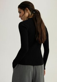 Czarna, prążkowana bluzka z długim rękawem o dopasowanym kroju, z subtelnym wysokim dekoltem i teksturowanym materiałem. W zestawieniu z szarymi, luźnymi spodniami.