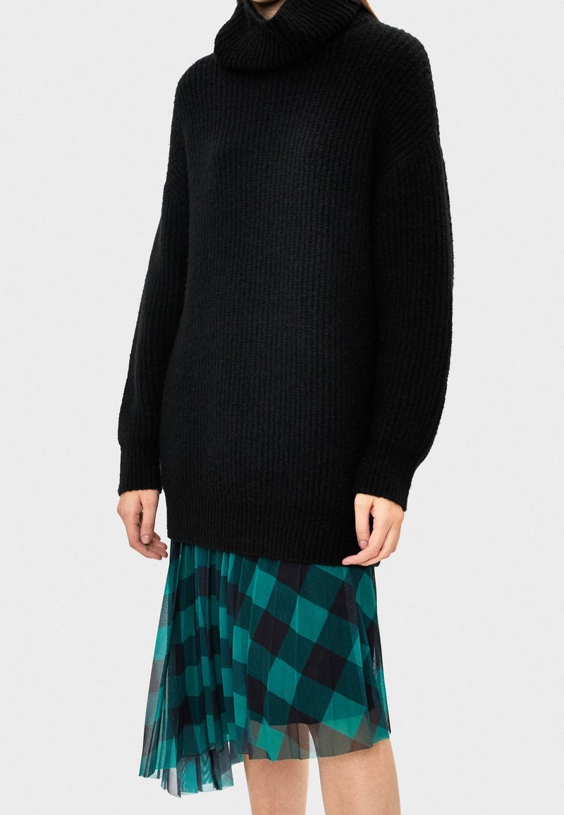 Maglione nero lavorato a maglia con colletto alto e texture a coste, abbinato a una gonna plissè in teal con motivo a rombi neri.