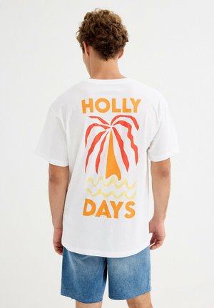 Hombre con camiseta blanca con estampado de palmera y olas en naranja y rojo y texto "HOLLY DAYS" en la espalda, combinado con pantalones cortos vaqueros azules.