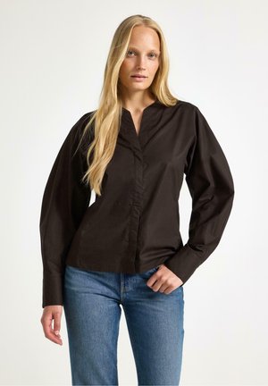 Lyshåret kvinde med langt hår iført en sort bluse med lange ærmer og blå jeans, stående mod en ensfarvet lys baggrund.