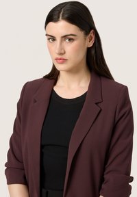 Burgundi ülikondjakk struktureeritud krae ja rullitud varrukatega, kantud musta soonilise topi üle. Sile kangas, mis sobib kehakujuga ning minimaalsete detailidega.