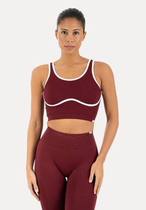 Kvinde stående, iført matchende burgunderfarvet sports-bh med hvide kanter og højtaljede leggings, vendt mod front mod ensfarvet baggrund.