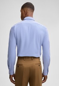 Camicia a maniche lunghe a righe blu chiaro con colletto button-down, vista da dietro, abbinata a pantaloni marroni. Texture del tessuto liscia.