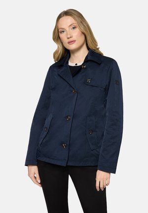 Kobieta ubrana w granatową kurtkę zapinaną na guziki z kołnierzem, stojąca na tle jasnego, jednolitego tła.