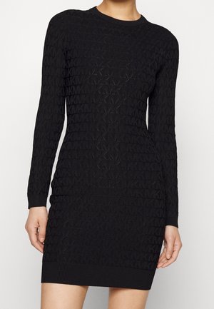 Mujer vistiendo un vestido de punto negro de manga larga con un patrón texturizado repetitivo de "MK", silueta ajustada y dobladillo y puños acanalados.