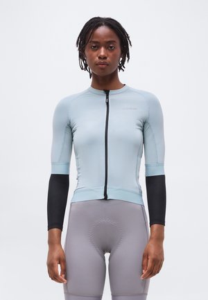 Mujer vistiendo un maillot de ciclismo ajustado de color azul claro con mangas negras y pantalones cortos de ciclismo acolchados grises, de pie contra un fondo liso.