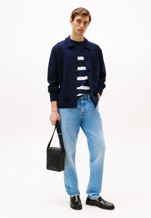Cardigan navy con bottoni, camicia a righe, jeans azzurri, scarpe in pelle nere e una piccola borsa a spalla nera. Sfondo bianco semplice.
