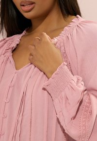 Blusa rosa testurizzata con scollatura e polsini a volant, bottoni frontali e dettagli in pizzo lungo i lati. Tessuto morbido con una vestibilità rilassata.