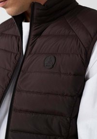 Bruine gewatteerde bodywarmer met hoge kraag, ritssluiting en logo op de borst. Heeft een doorgestikt ontwerp en een gladde textuur. Gedragen over een wit shirt.