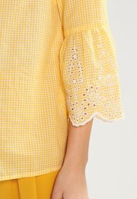 Gul gingham-blus med trumpetärmar. Ögla spetskant vid ärmsluten ger textur. Tyget är lätt, med rutigt mönster genom hela.