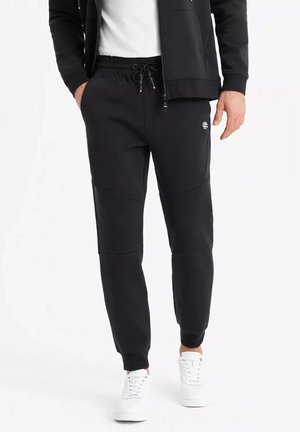 Pantalons de survêtement noirs en tissu doux avec une taille élastique, des poches latérales et un détail de logo près de la cuisse. Dispose d'un design de jambe fuselée.