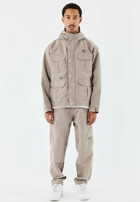 Chaqueta impermeable beige con capucha, que presenta múltiples bolsillos frontales y cierres de botón a presión, combinada con pantalones cargo a juego y zapatillas blancas.