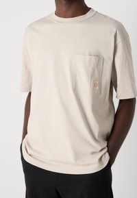 Beige bomulls t-shirt med korta ärmar, rund halsringning och en vänster bröstficka med en patch-logga. Mjuk textur och avslappnad passform.