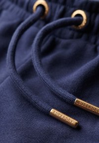 Navyblauwe sweatpants met twee trekkoorden, voorzien van gouden metalen uiteinden met de gravure "SUPERDRY." Zachte, gestructureerde stof.