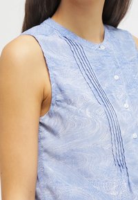 Blusa senza maniche azzurro chiaro con un motivo testurizzato a spirale, bottoniera con dettagli arricciati e bottoni bianchi lungo il davanti.
