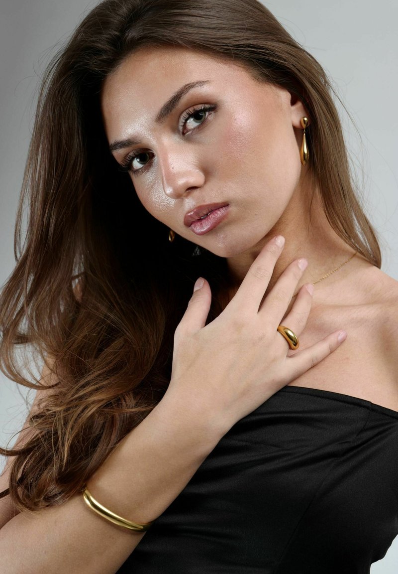 Joyería de oro, incluyendo pendientes de aro, un anillo y una pulsera, sobre una blusa negra texturizada. Una luz suave y cálida realza el acabado metálico.