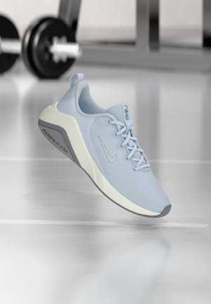 Chaussure de course Nike Air Zoom bleu clair flottant avec des poids de gym flous en arrière-plan sur un sol réfléchissant.
