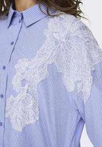 Camisa abotoada em azul claro com riscas verticais brancas, apresentando um intrincado aplique de renda branca no ombro esquerdo.