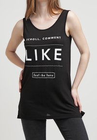 Svart ärmlös linne med fet vit text: "SKROLLA, KOMMENTERA, GILLA" och "känn kärleken." Mjuk tyg med en avslappnad passform.