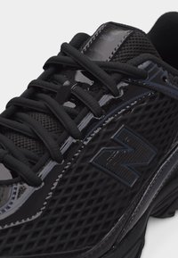 Κοντινό πλάνο μαύρου παπουτσιού New Balance με υφή από δίχτυ, γυαλιστερές επενδύσεις και χοντρές κορδέλες σε λευκό φόντο.