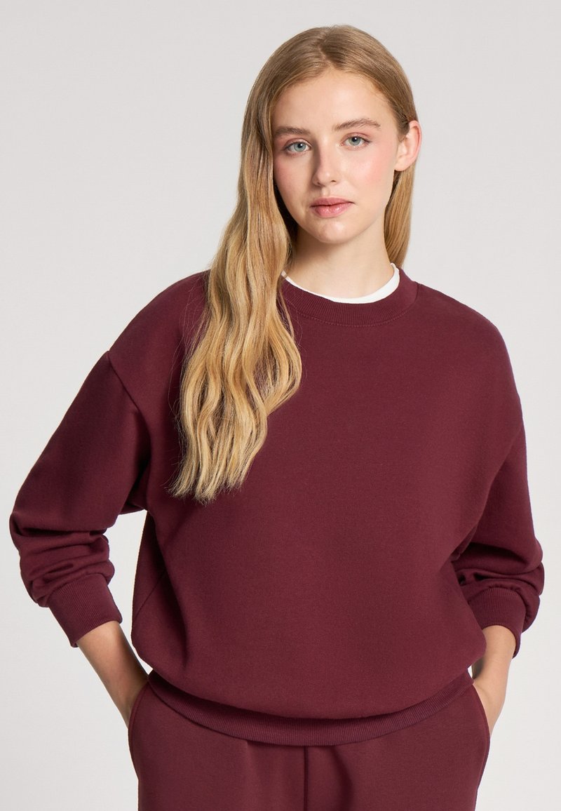 Burgundowy sweatshirt z okrągłym dekoltem, opadającymi ramionami i prążkowanymi mankietami. Miękka tekstura, casualowy design, w zestawie z dopasowanymi spodniami.