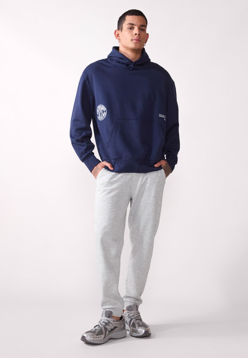 Marineblauer Hoodie mit Fronttasche und Logodetail, kombiniert mit hellgrauen Jogginghosen. Abgerundet mit grauen Sportschuhen.