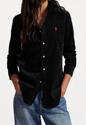 Femme portant une chemise en velours côtelé noire avec un logo rouge sur la poitrine, boutonnée, et un jean bleu, les mains dans les poches.
