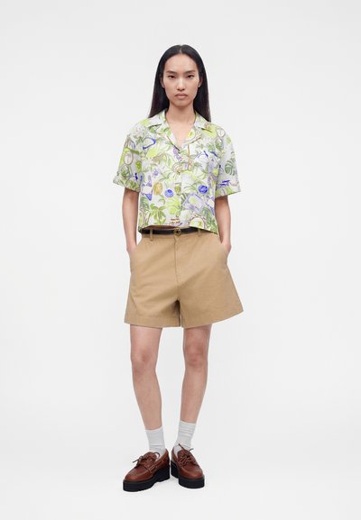 Γυναίκα στέκεται με το πρόσωπο προς τα εμπρός φορώντας ένα κοντομάνικο πουκάμισο με λουλούδια και κουμπιά, μπεζ σορτς με ψηλή μέση, λευκές κάλτσες και καφέ παπούτσια με πλατφόρμα.