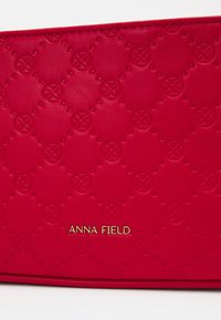 Portefeuille en cuir rouge avec un motif texturé de petits cercles et croix, arborant un logo doré "ANNA FIELD" en bas.