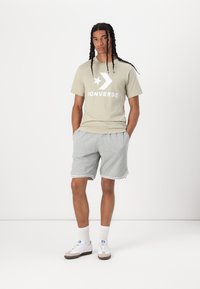 Gai olīvu t-kreklu ar baltu Converse logo, apvienojot to ar pelēkiem sporta šortiem, kuriem ir balta apmale, un papildinot ar baltām un pelēkām sporta kurpēm.