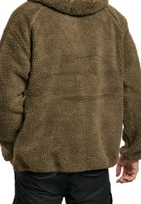 Giacca in pile marrone con superficie testurizzata e cappuccio. Presenta una vestibilità ampia e polsini elasticizzati per calore e comfort.